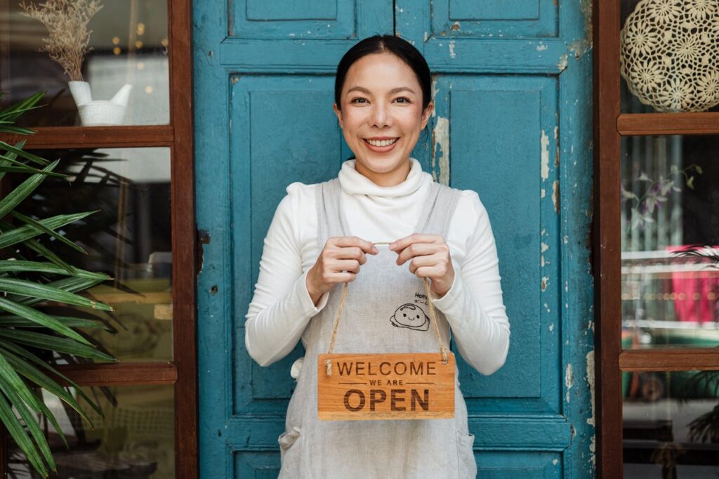 ethnic-female-cafe-owner-showing-welcome-we-are-open-inscription-4473398 Cheerful ethnic female cafeteria owner in apron demonstrating cardboard signboard while standing near blue shabby door and windows after starting own business and looking at camera